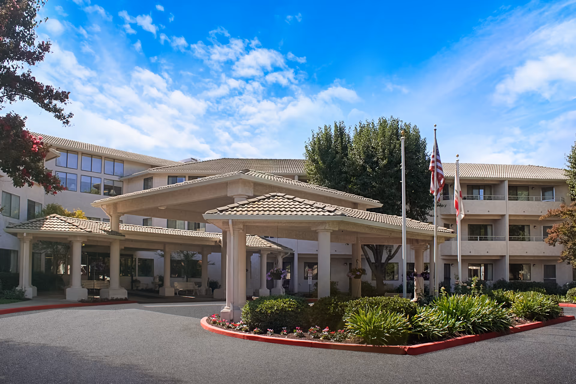 Front entrance of a multi-story senior living facility with a covered porte-cochere, flagpoles, and landscaped driveway under a blue sky.