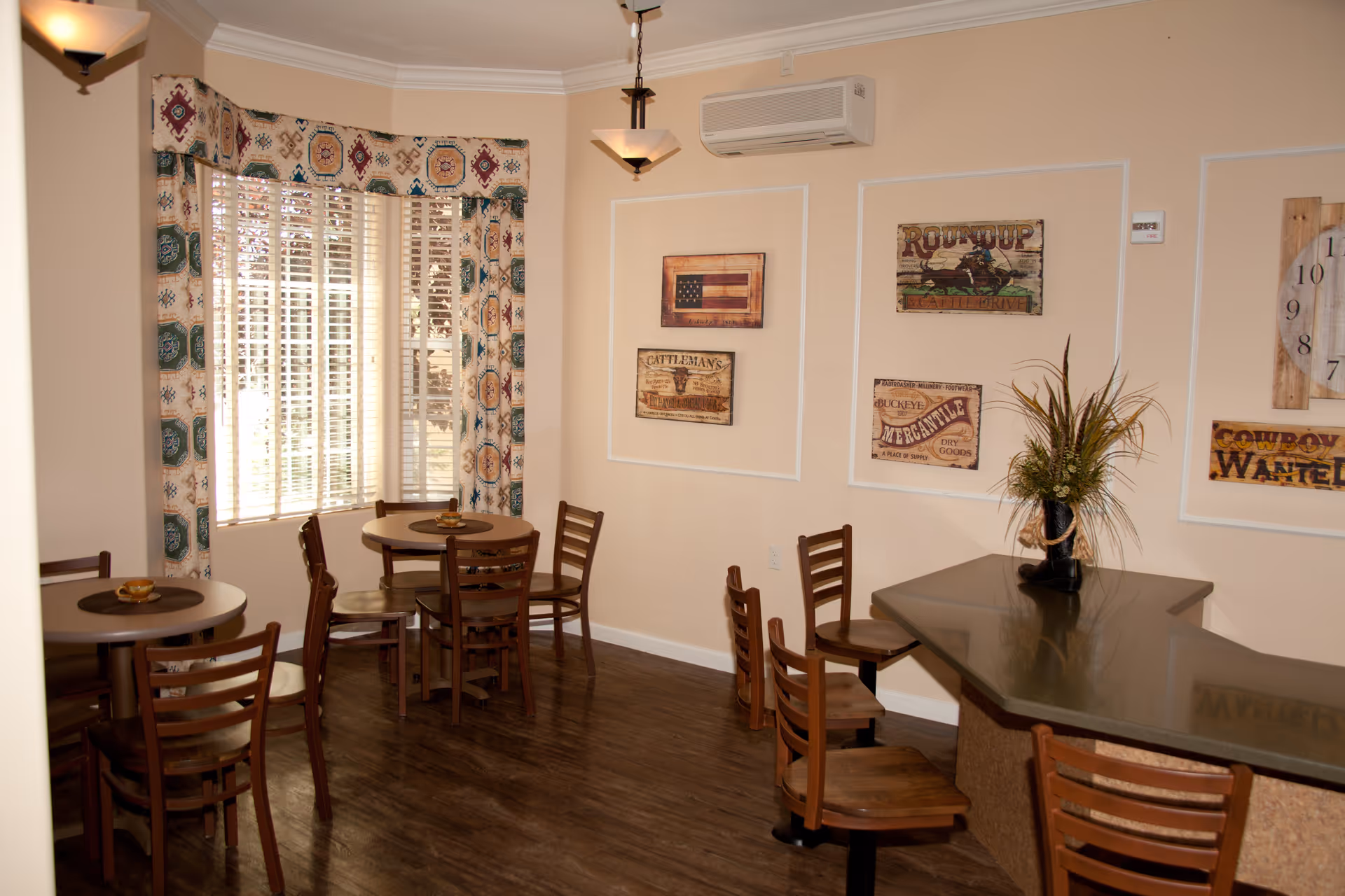 A cozy dining area with round wooden tables and chairs, a large window with patterned curtains, and wall decorations including rustic signs and a clock. There is a counter with additional seating and a decorative plant in a boot-shaped vase.