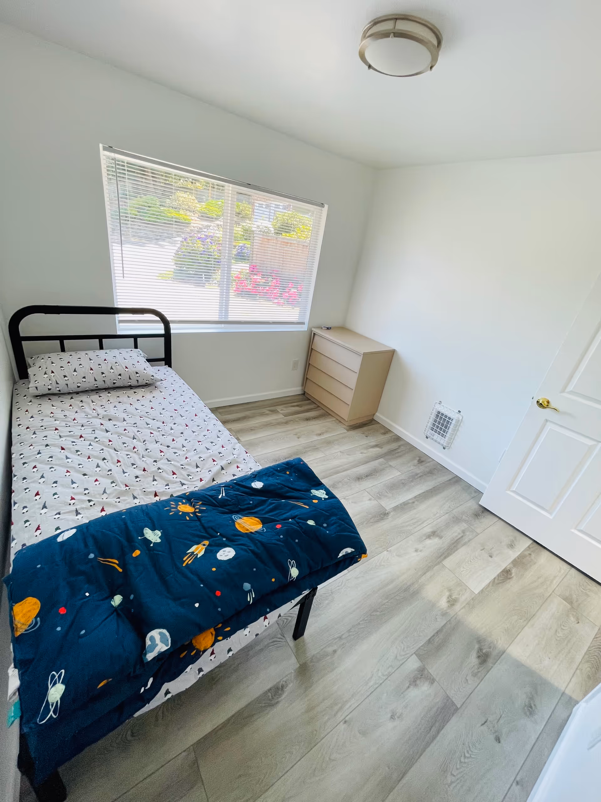 A small, bright bedroom with a single bed featuring space-themed bedding and a pillow with a pattern of small figures. There is a window with blinds letting in natural light, a light wood dresser against the wall, light-colored wood flooring, and a white door with a gold handle.