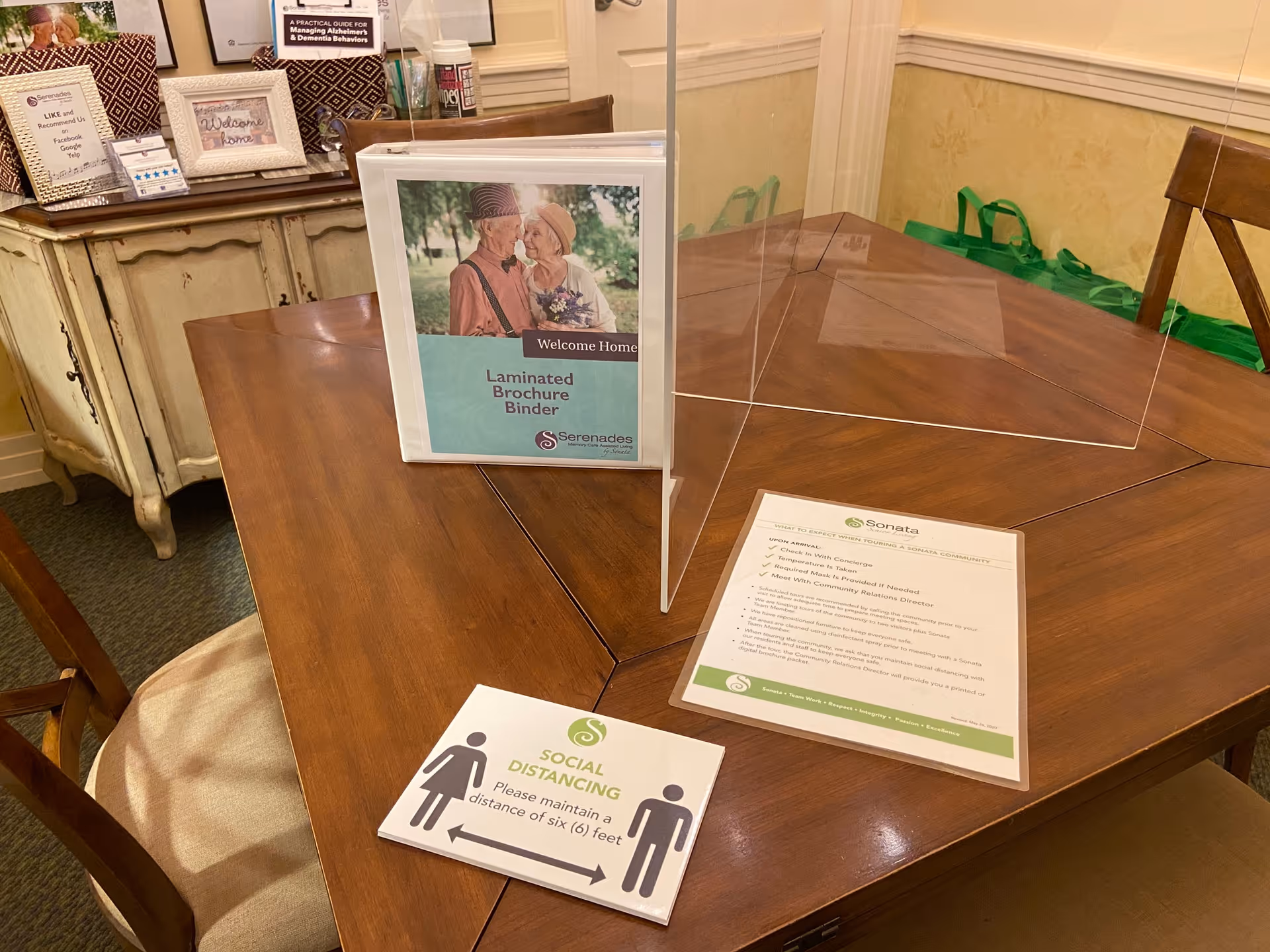 A wooden table in a senior living facility with a laminated brochure binder, social distancing sign, paperwork, chairs, and a plexiglass divider.
