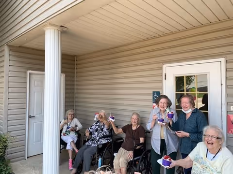 A group of six elderly women sitting and standing outside a building with beige siding, smiling and holding cups of ice cream or dessert. Two women are in wheelchairs, and the others are standing or sitting on chairs near a white door and a white column.