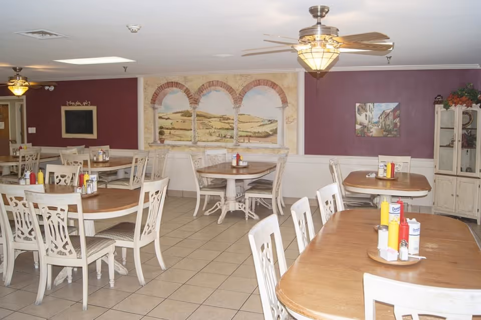 Dining room with multiple wooden tables and white chairs, condiments on the tables, a mural on the wall and ceiling fans overhead.