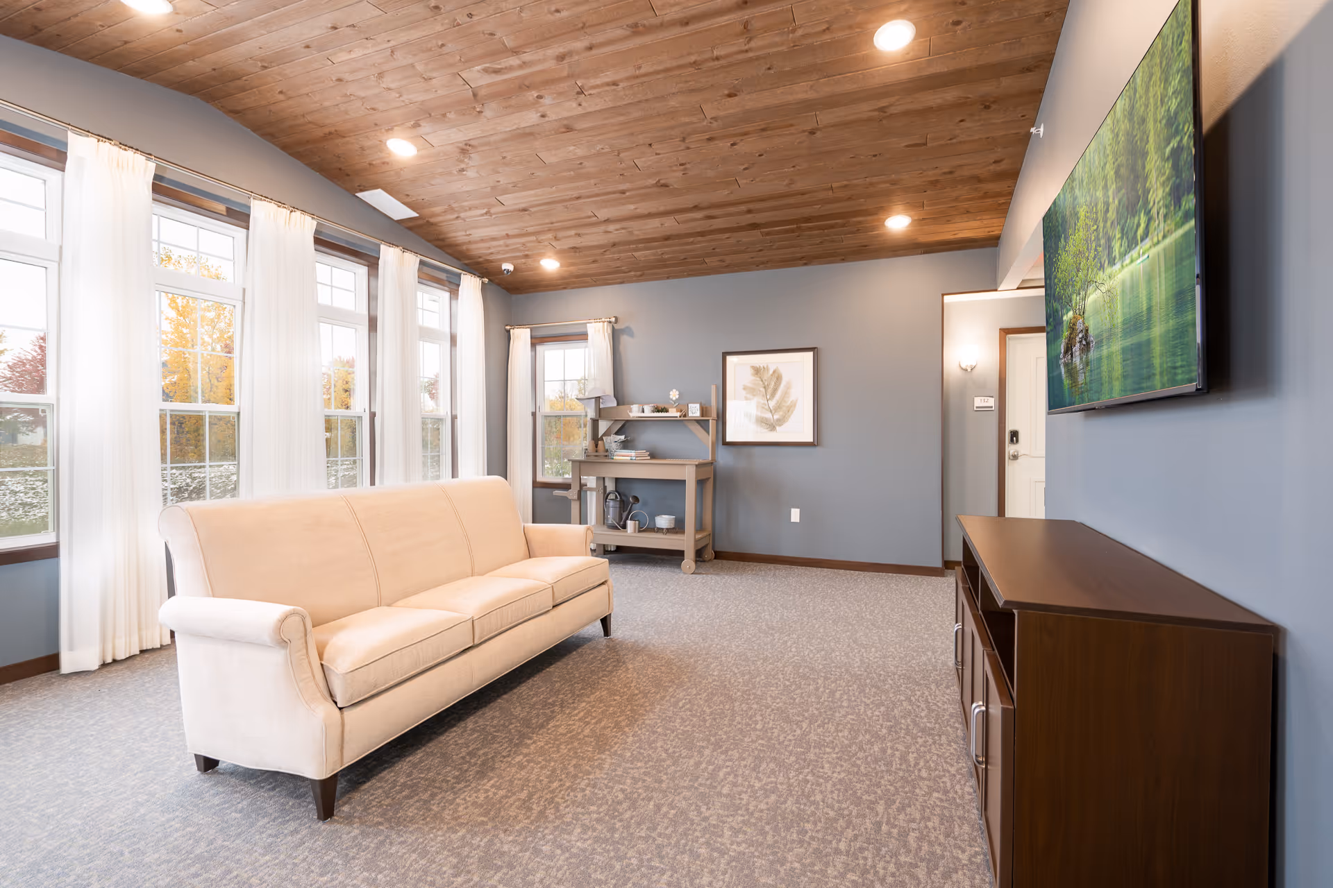 A bright living room with a beige sofa facing a wall-mounted flat screen TV. The room features large windows with white curtains, a wooden ceiling, gray walls, and a carpeted floor. There is a small wooden shelf with decorative items and a framed leaf artwork on the wall.