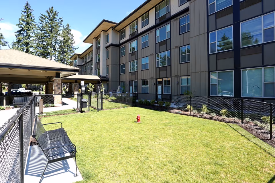 Outdoor area of a senior living facility with a fenced grassy lawn, a black metal bench on a concrete path, and a multi-story building in the background under a clear blue sky.