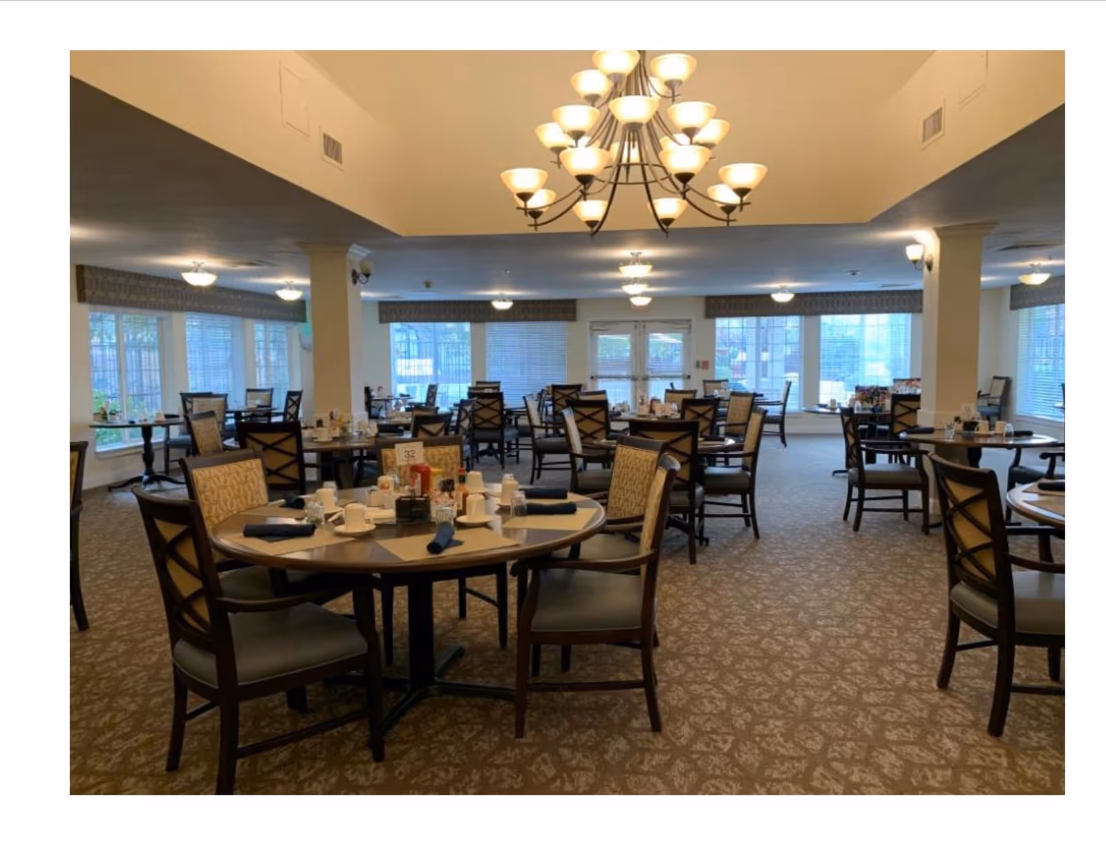 Dining room with multiple round tables set for meals, upholstered chairs, chandeliers, and large windows.