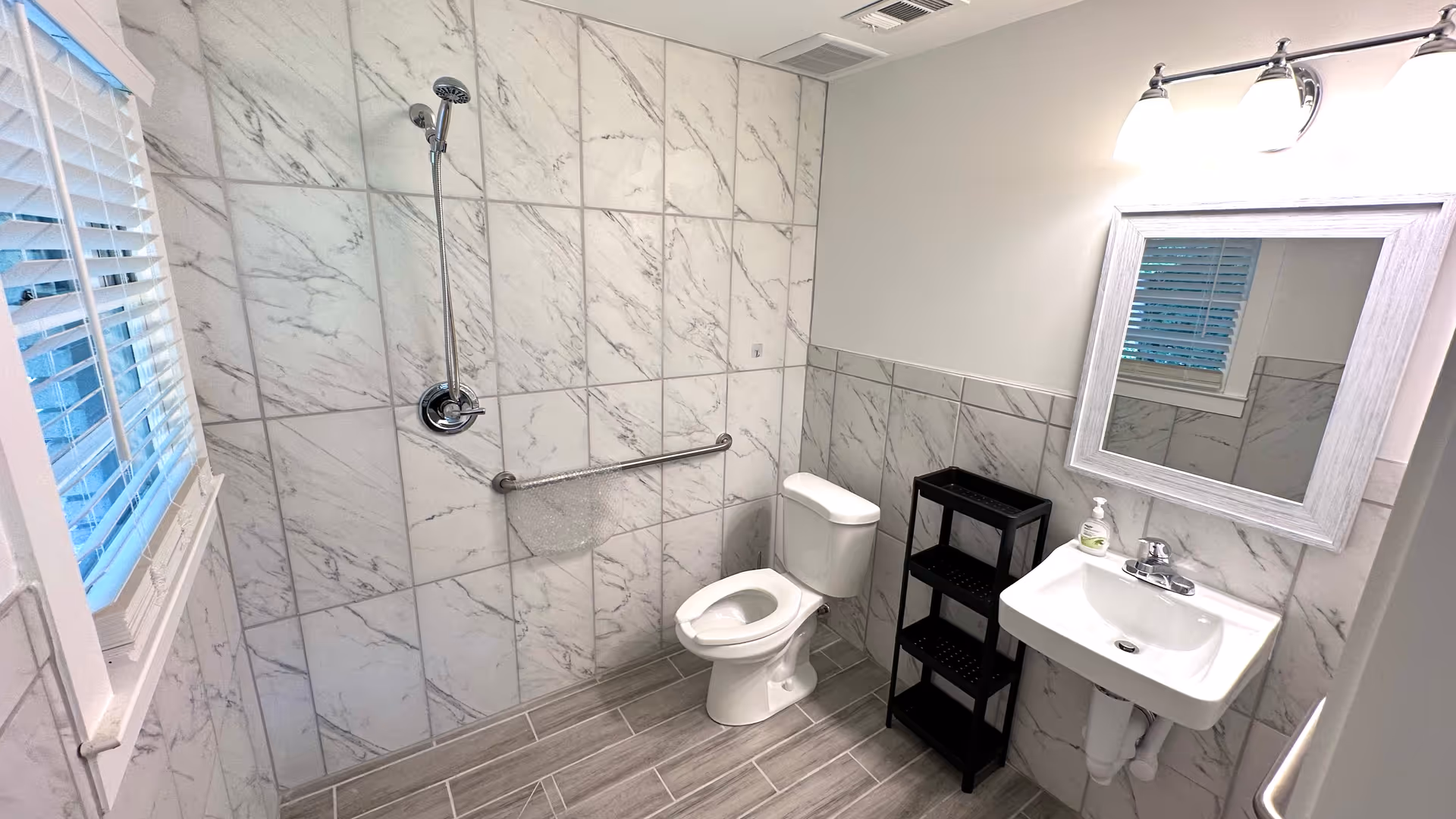 A clean and modern senior living bathroom with white marble-patterned tiles on the walls and gray floor tiles. The bathroom features a walk-in shower with a handheld showerhead and a grab bar, a white toilet, a white sink with a mirror above it, a black three-tier shelf, and a window with white blinds.