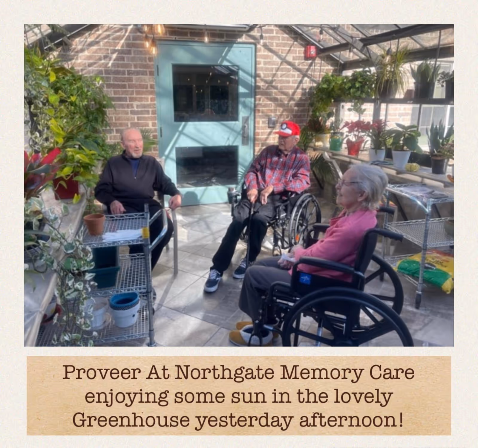 Three elderly individuals, two men and one woman, sitting in wheelchairs inside a sunlit greenhouse with brick walls and a glass ceiling. The greenhouse is filled with various potted plants on shelves and tables. One man is wearing a red cap and a plaid shirt, while the woman is dressed in a pink top. They appear to be enjoying the sunlight and the greenery around them.