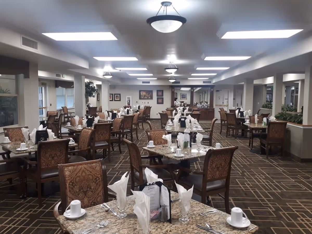 A spacious dining room with multiple tables set with white napkins, cups, and silverware. The room has patterned carpet flooring, cushioned chairs with ornate backs, and bright overhead lighting. There are plants and framed artwork on the walls, creating a welcoming atmosphere.