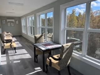 Sunlit interior corridor with paired tables and upholstered chairs arranged along a row of large windows overlooking trees.