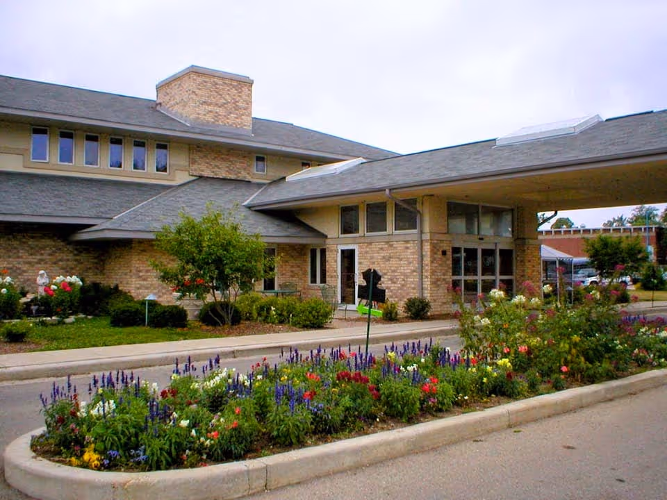 Exterior view of a brick building with a covered entrance and a landscaped flower bed in front, featuring various colorful flowers and greenery under an overcast sky.