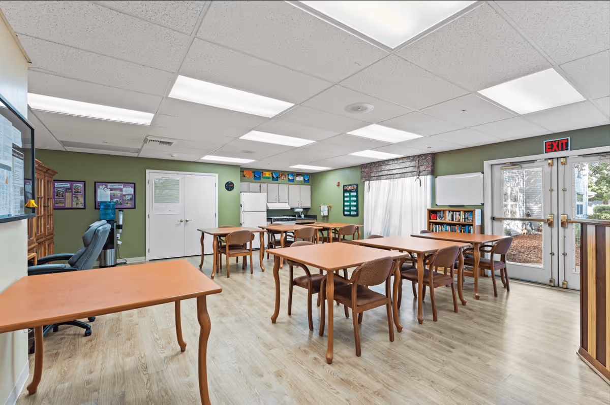 A well-lit room with several wooden tables and chairs arranged neatly on a light wood floor. The walls are painted green, and there is a kitchen area with white cabinets, a refrigerator, and a stove in the background. A water cooler, bulletin boards, and a bookshelf are also visible. Double glass doors with an exit sign above them lead outside.