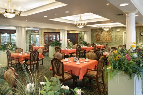 A spacious dining room with multiple tables covered in coral-colored tablecloths, each set with white napkins and silverware. The room features elegant wooden chairs with patterned upholstery, floral arrangements on some tables, and decorative plants in the foreground. The walls are adorned with wallpaper and a large tapestry, and the ceiling has recessed lighting and chandeliers. Large windows allow natural light to enter the room.