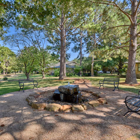 Outdoor area at Kruse Village featuring a circular stone fountain surrounded by gravel and several black metal benches. Tall trees provide shade and greenery, with a building visible in the background under a clear blue sky.
