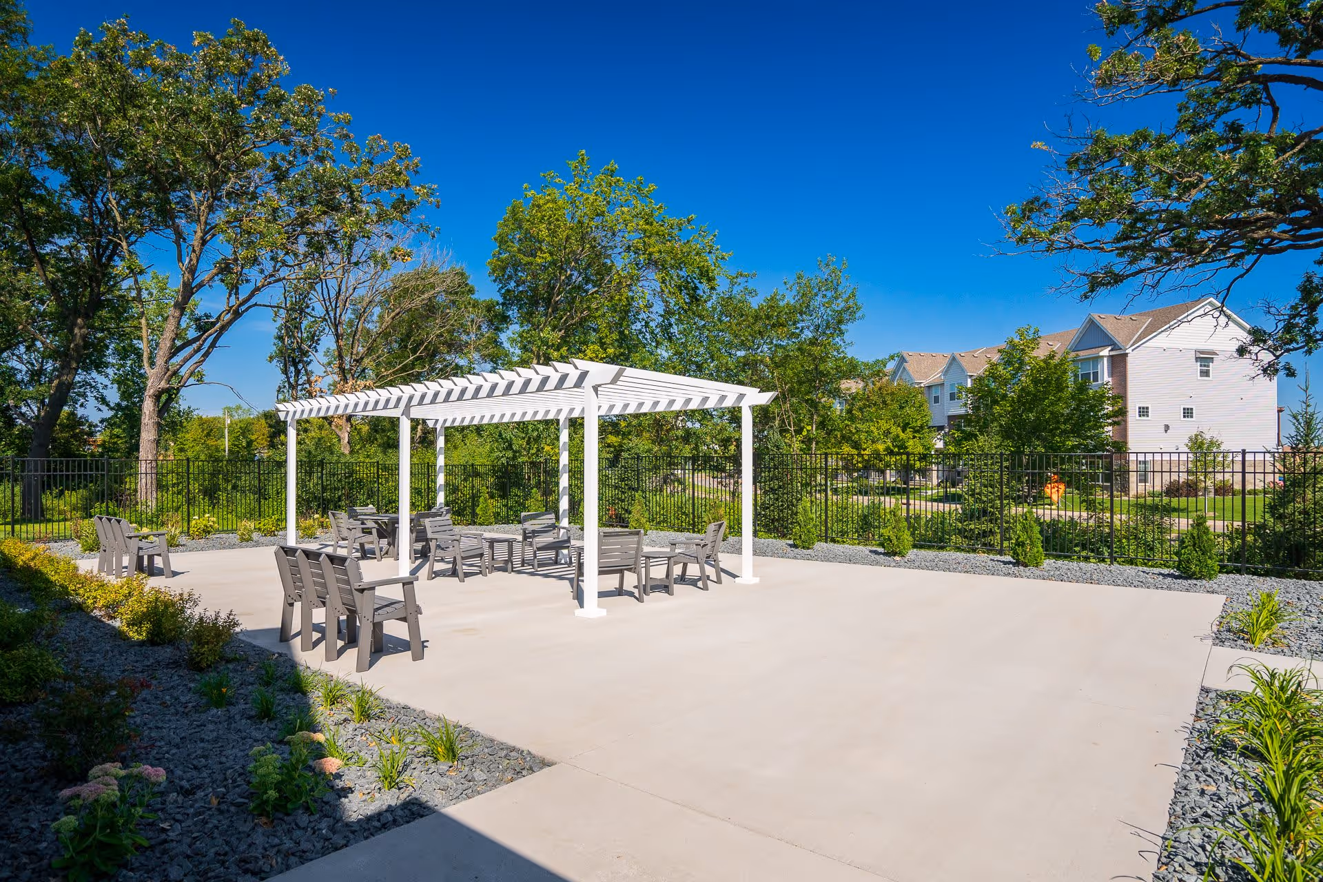 Outdoor patio area with a white pergola providing shade over several gray chairs and tables. The patio is surrounded by a black metal fence, greenery, and trees under a clear blue sky. Residential buildings are visible in the background.