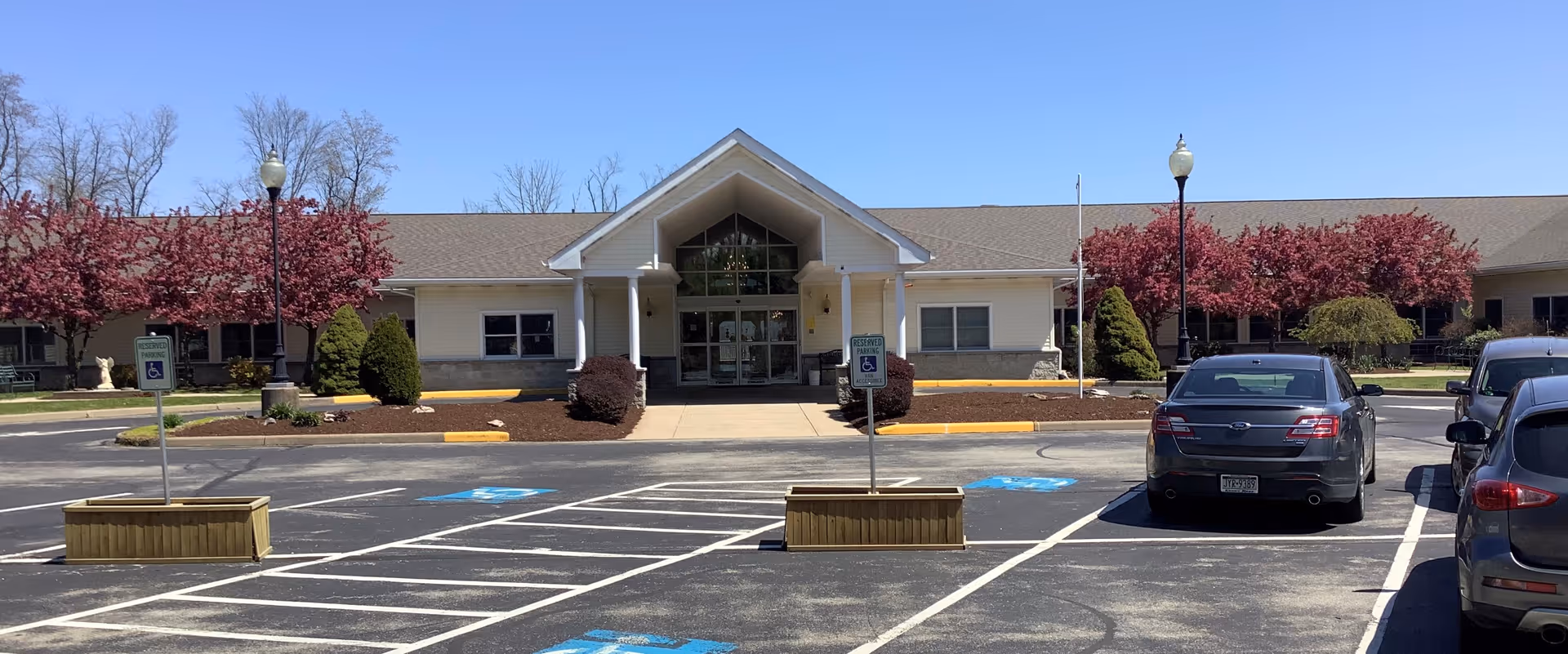Front exterior view of Golden Heights Personal Care facility with a parking lot in front, including several handicap parking spaces. The building has a peaked roof entrance with glass doors and windows, surrounded by landscaped bushes and trees with pink blossoms under a clear blue sky.