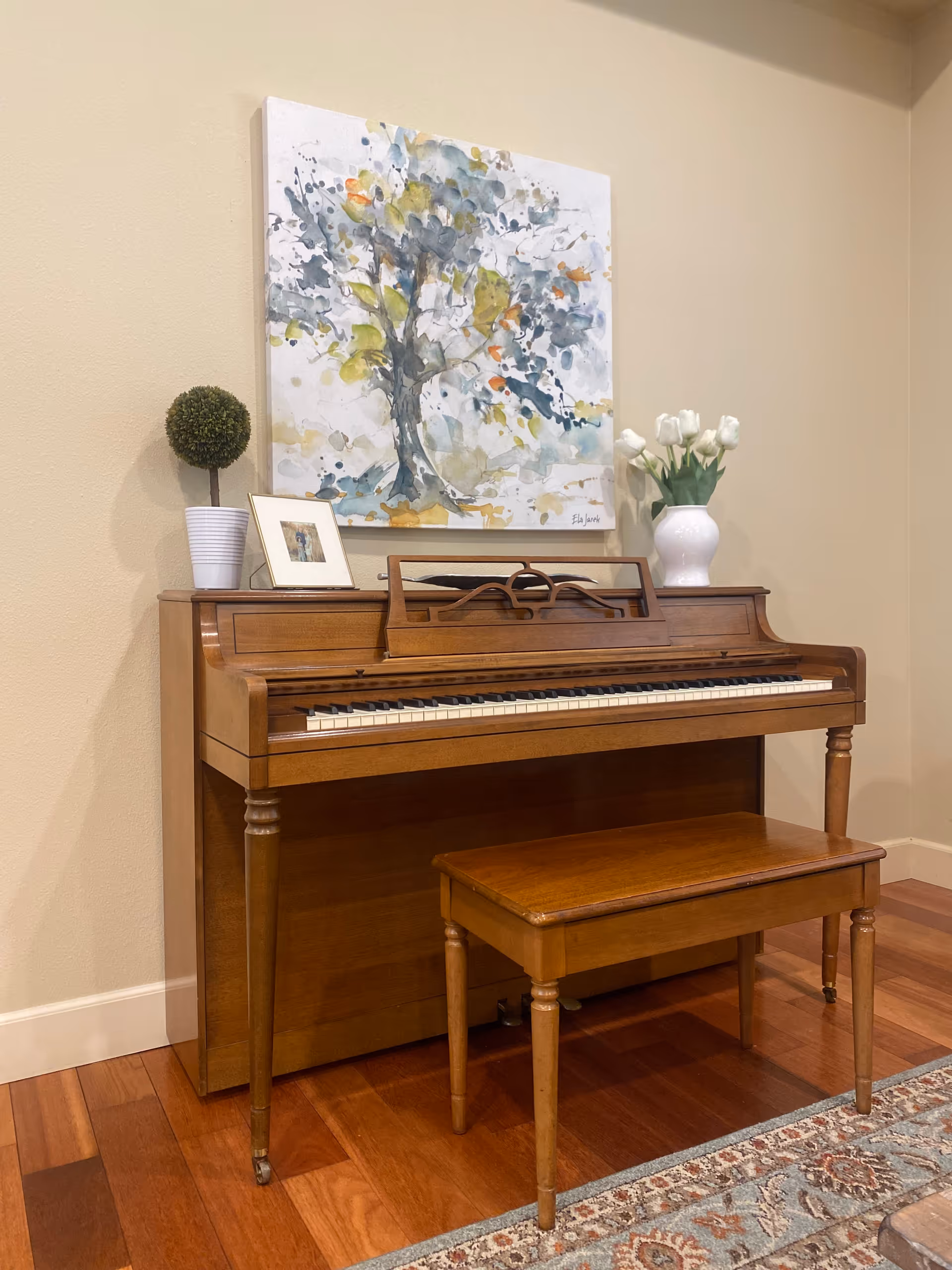 A wooden upright piano with a matching bench in a room with hardwood floors. On top of the piano are a small potted plant, a framed photo, and a white vase with white tulips. Above the piano hangs a colorful abstract painting of a tree.