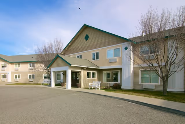 Exterior view of a two-story senior living facility building with beige siding and green trim. The entrance has a covered porch with white columns and a white bench nearby. Leafless trees are visible around the building under a clear sky.