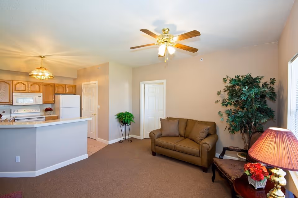 Interior view of a living area and kitchen in a senior living facility. The living area features a brown loveseat with two cushions, a side table with a lamp and a flower arrangement, and a large potted plant in the corner. The kitchen has wooden cabinets, a white refrigerator, a white microwave, and a white stove. A ceiling fan with lights is mounted on the ceiling, and the walls are painted beige.