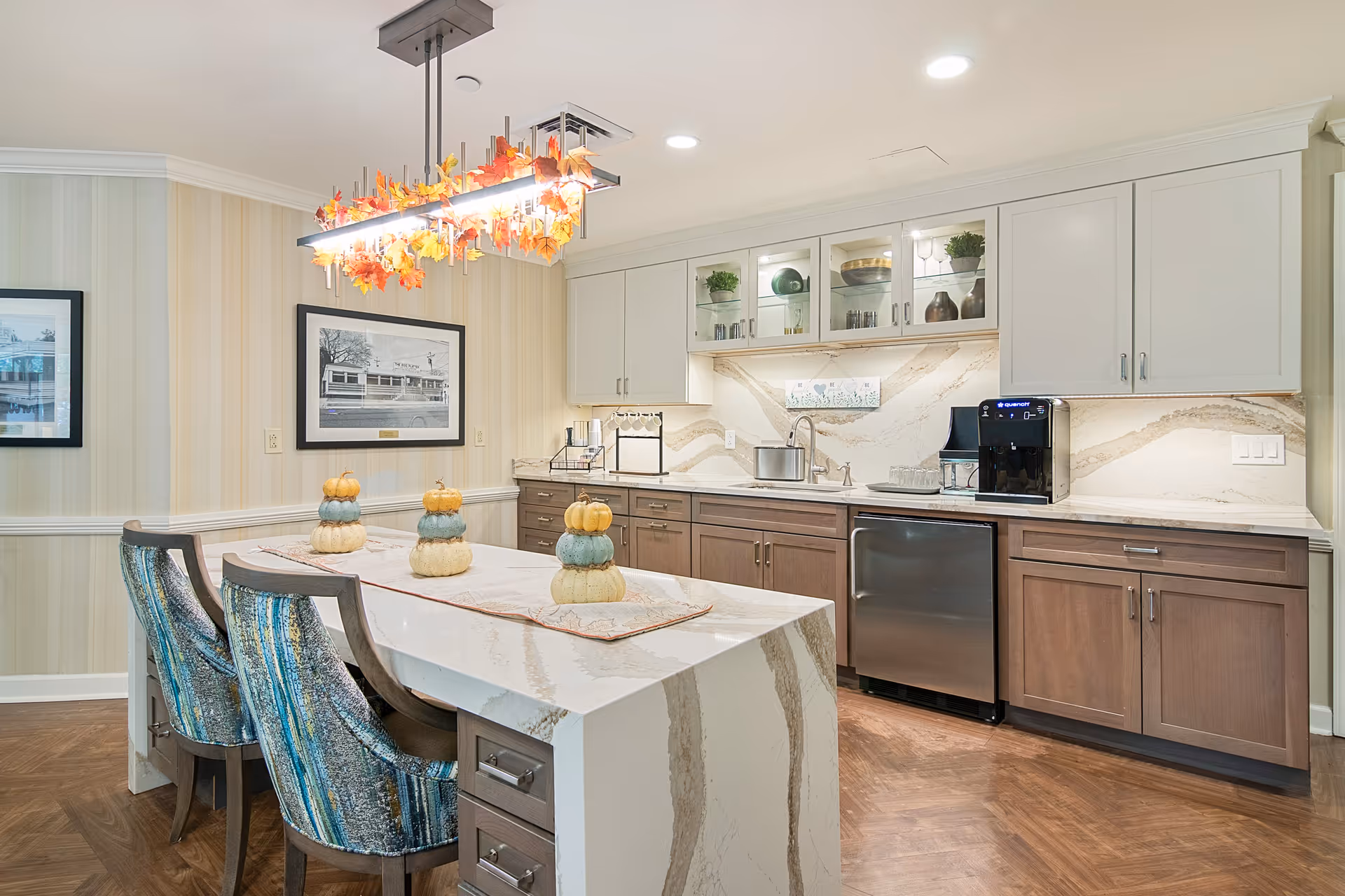 A modern kitchen area with a large marble island countertop decorated with three stacked pumpkins. The kitchen features wooden lower cabinets, white upper cabinets with glass doors displaying decorative items, a stainless steel dishwasher, and a coffee machine. Above the island hangs a rectangular light fixture adorned with autumn leaves. The walls have light-colored striped wallpaper and framed black-and-white photos.