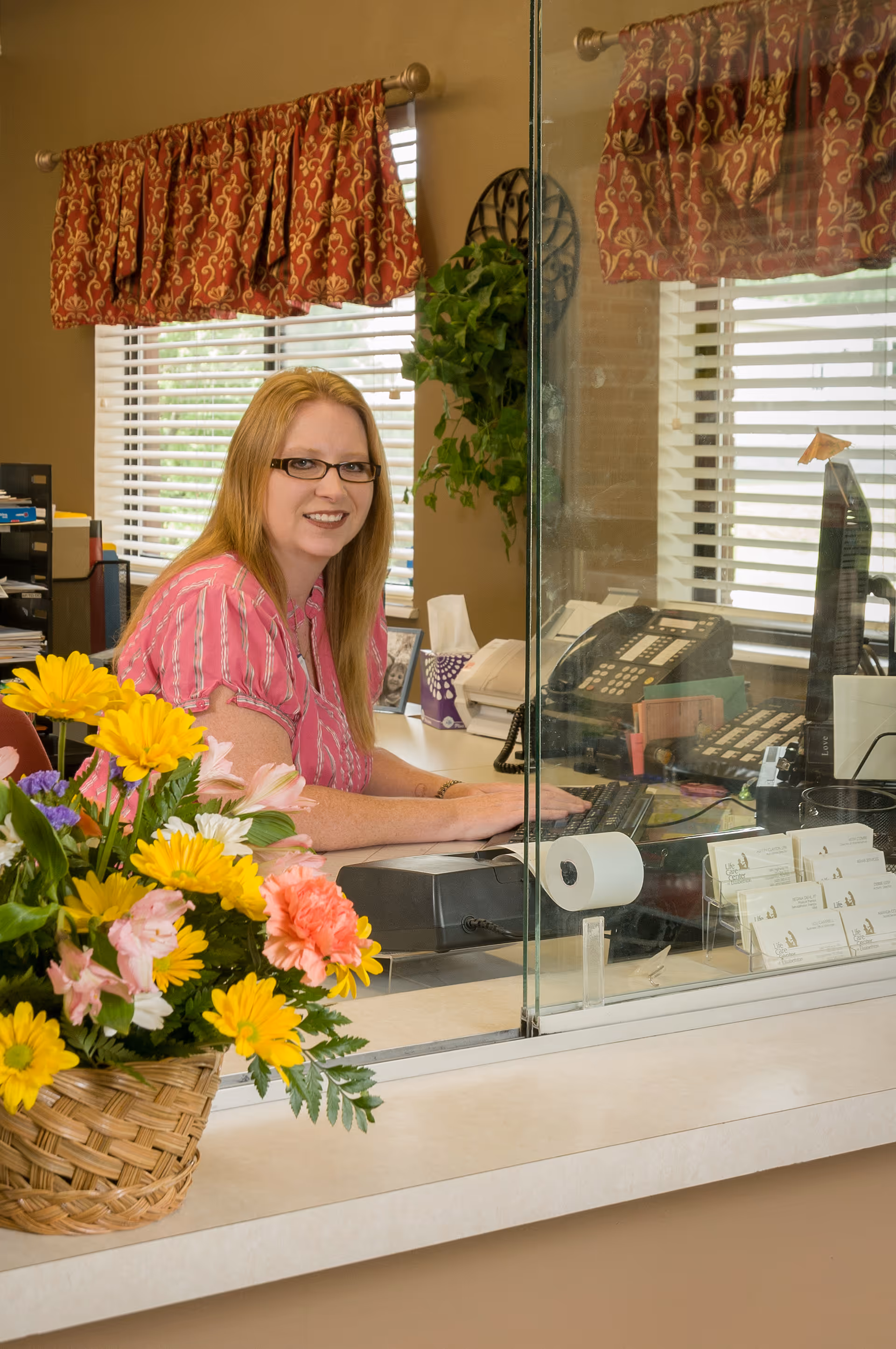 A woman with glasses and long blonde hair wearing a pink striped shirt is sitting behind a glass partition at a reception desk. There is a basket of colorful flowers on the counter in front of her, and office equipment including a telephone and computer keyboard is visible behind the glass. The background shows a window with blinds and a red patterned valance, along with a green plant on the wall.