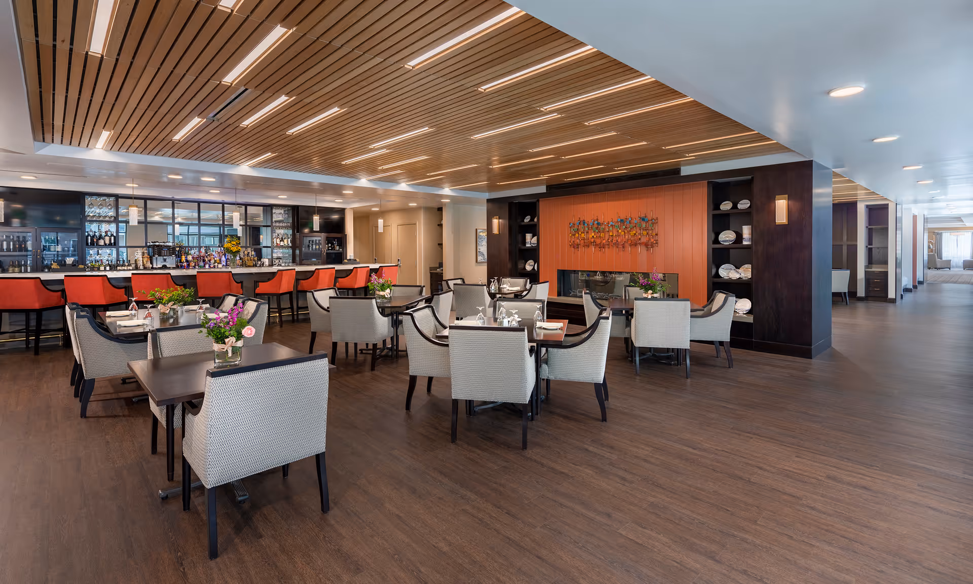 Spacious senior living dining room with tables and upholstered chairs, a bar with red stools, and a modern wood-slat ceiling.