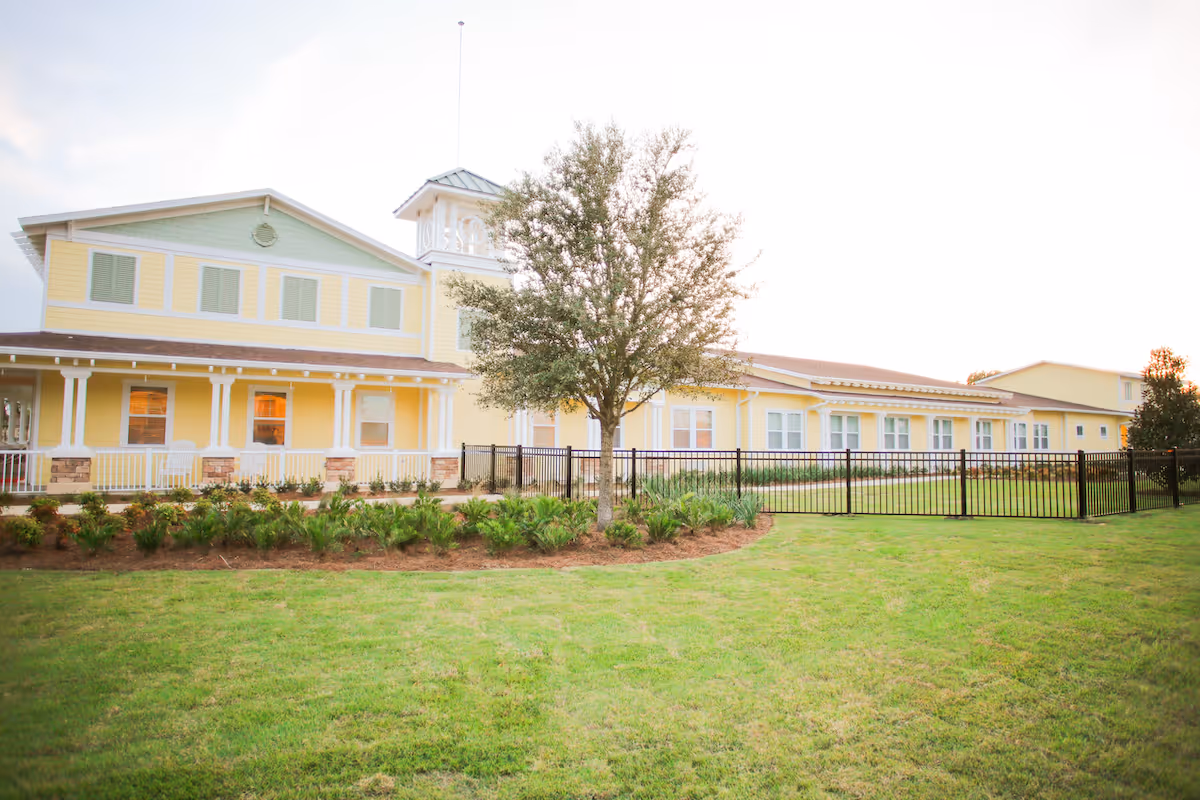 Yellow two-story building with a covered porch, a central tree, black metal fence, and a manicured lawn.