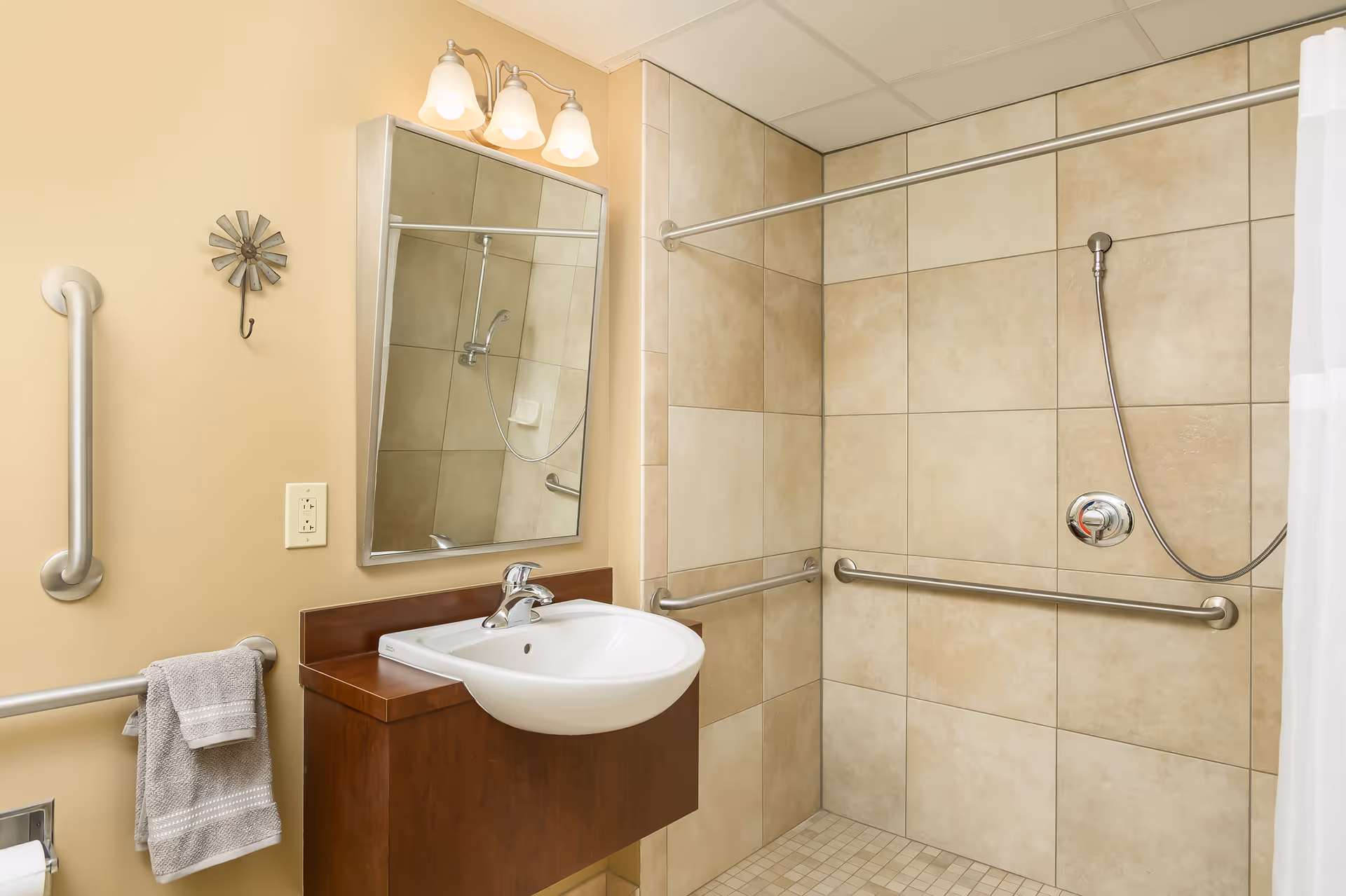 A bathroom with beige tiled walls and floor, featuring a walk-in shower with a handheld showerhead and grab bars. There is a white sink mounted on a wooden vanity with a mirror above it, and a towel hanging on a grab bar next to the sink. The walls are painted light beige and there is a three-light fixture above the mirror.