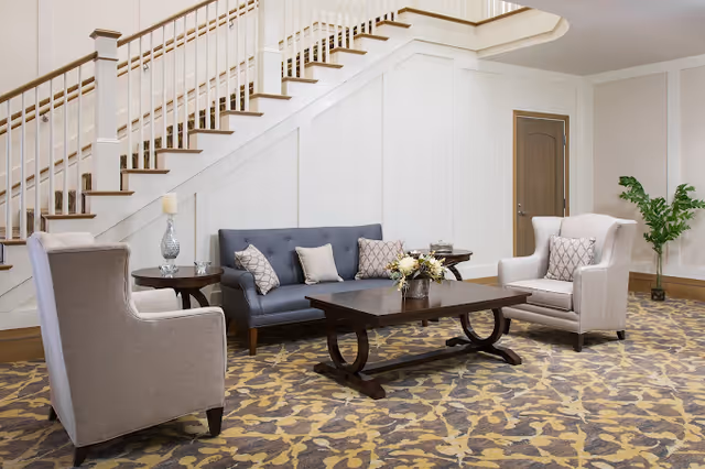 A cozy sitting area with a blue sofa, two beige armchairs, a dark wooden coffee table with a floral centerpiece, and a side table with a decorative lamp. The space is carpeted with a patterned rug and features a staircase with wooden handrails and white panel walls.