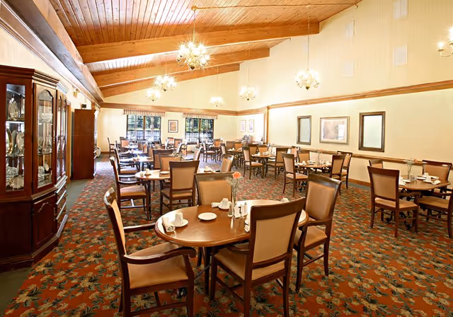 Spacious dining room with multiple round and rectangular tables and upholstered chairs beneath chandeliers and a wood-beamed ceiling.