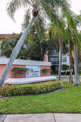 Exterior view of Elison Independent Living of Lake Worth facility with a sign displaying the name. The building is surrounded by palm trees, green grass, and shrubs under a clear sky.