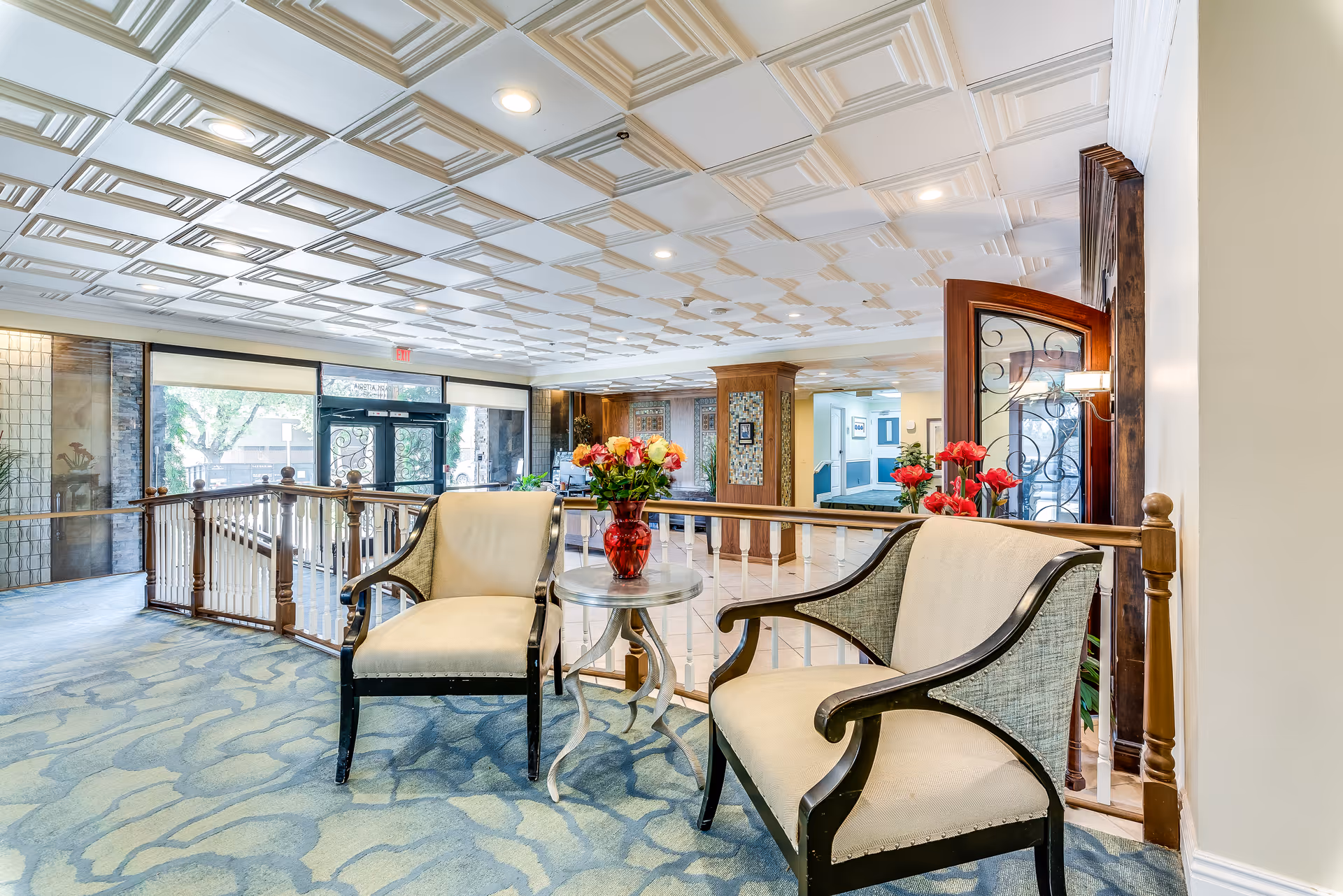 A bright and spacious senior living facility common area with two beige armchairs and a small round table with a red vase of flowers between them. The area features a patterned carpet, decorative ceiling tiles, wooden railing, and large windows letting in natural light.