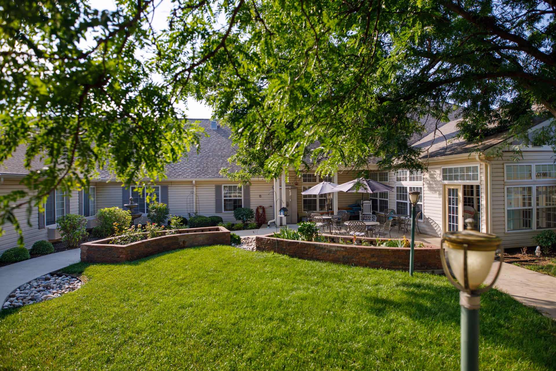 Grassy courtyard with patio seating and umbrellas surrounded by a single-story assisted living building and overhanging tree branches.