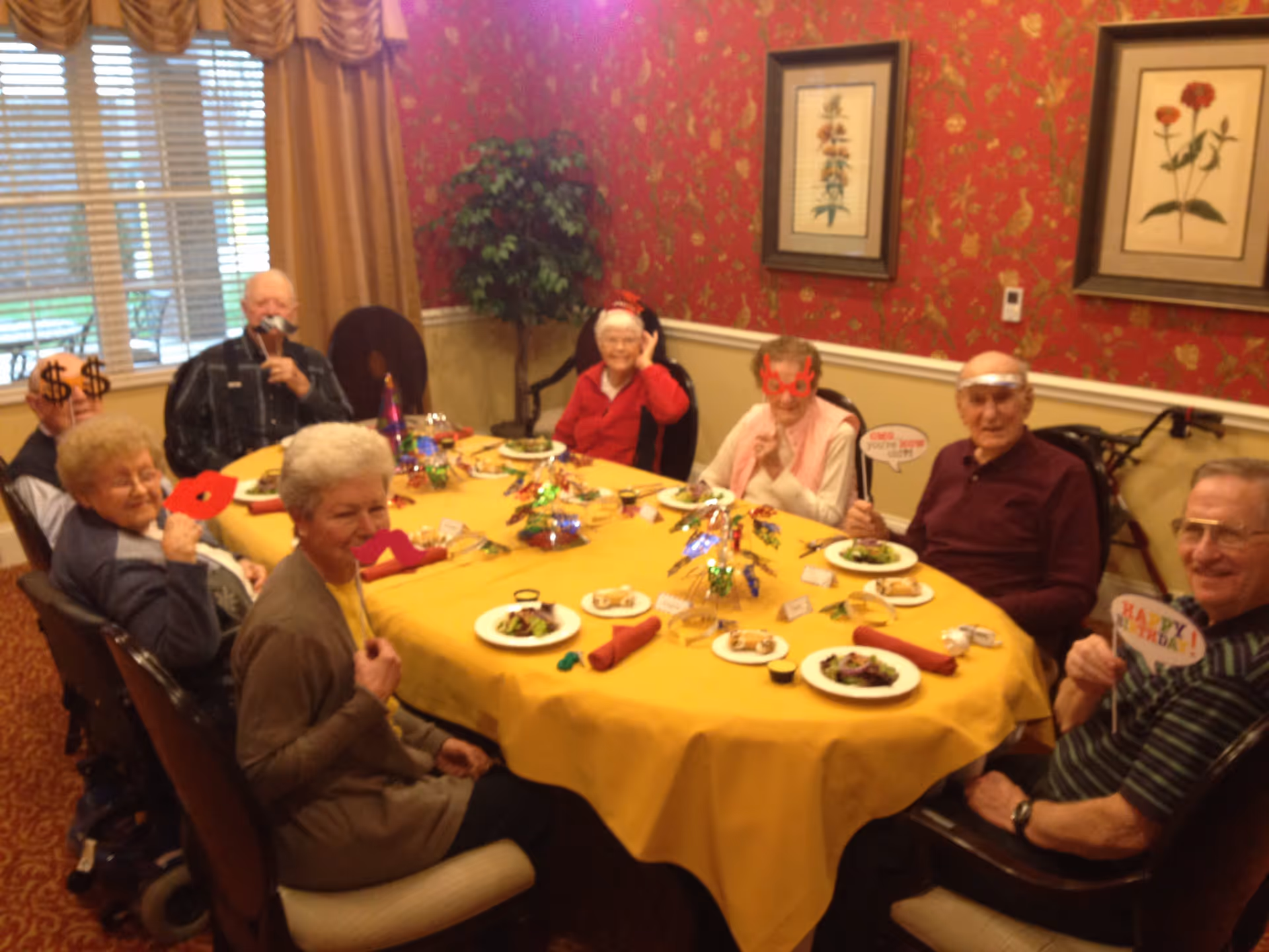 A group of elderly people sitting around a dining table with a yellow tablecloth, enjoying a meal together in a decorated room with red floral wallpaper and framed botanical prints. Some individuals are holding playful photo props like glasses, lips, and signs, suggesting a festive or celebratory atmosphere.