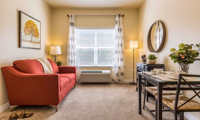 Bright living room with a red loveseat, window with patterned curtains, a small dining table, and decorative accents.