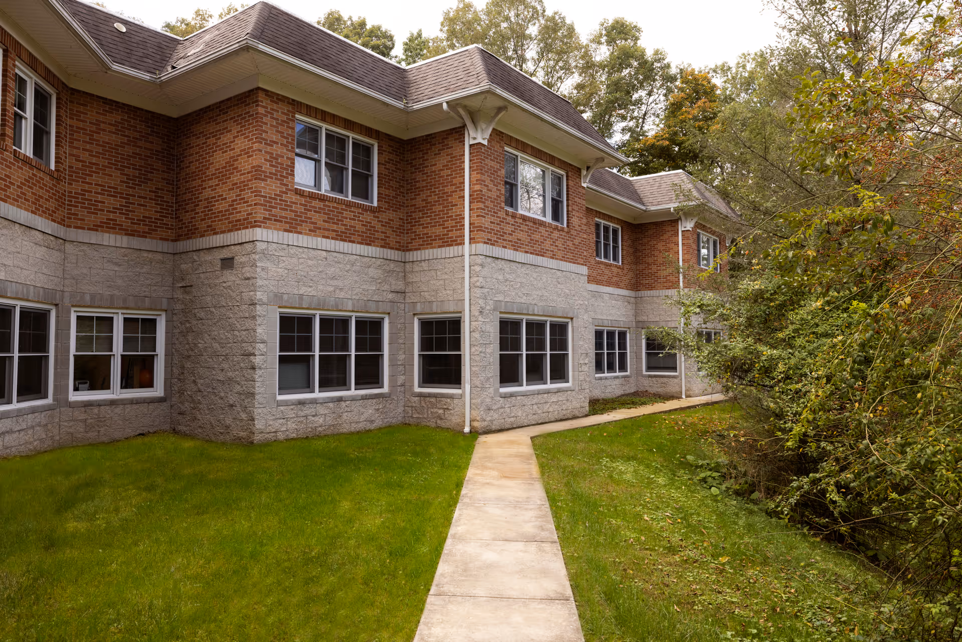 Exterior view of a two-story senior care facility building with brick and stone facade, multiple windows, a concrete walkway, green grass, and surrounding trees.