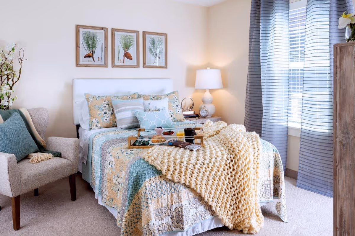 A cozy bedroom with a neatly made bed featuring a patchwork quilt and multiple pillows. A chunky knit blanket is draped over the bed, which also has a wooden tray with breakfast items. To the left of the bed is a beige armchair with a blue cushion and a throw blanket. On the right side, there is a wooden nightstand with a white lamp, clock, and decorative items. The room has light-colored walls, three framed botanical prints above the bed, and a window with sheer striped curtains allowing natural light to enter.