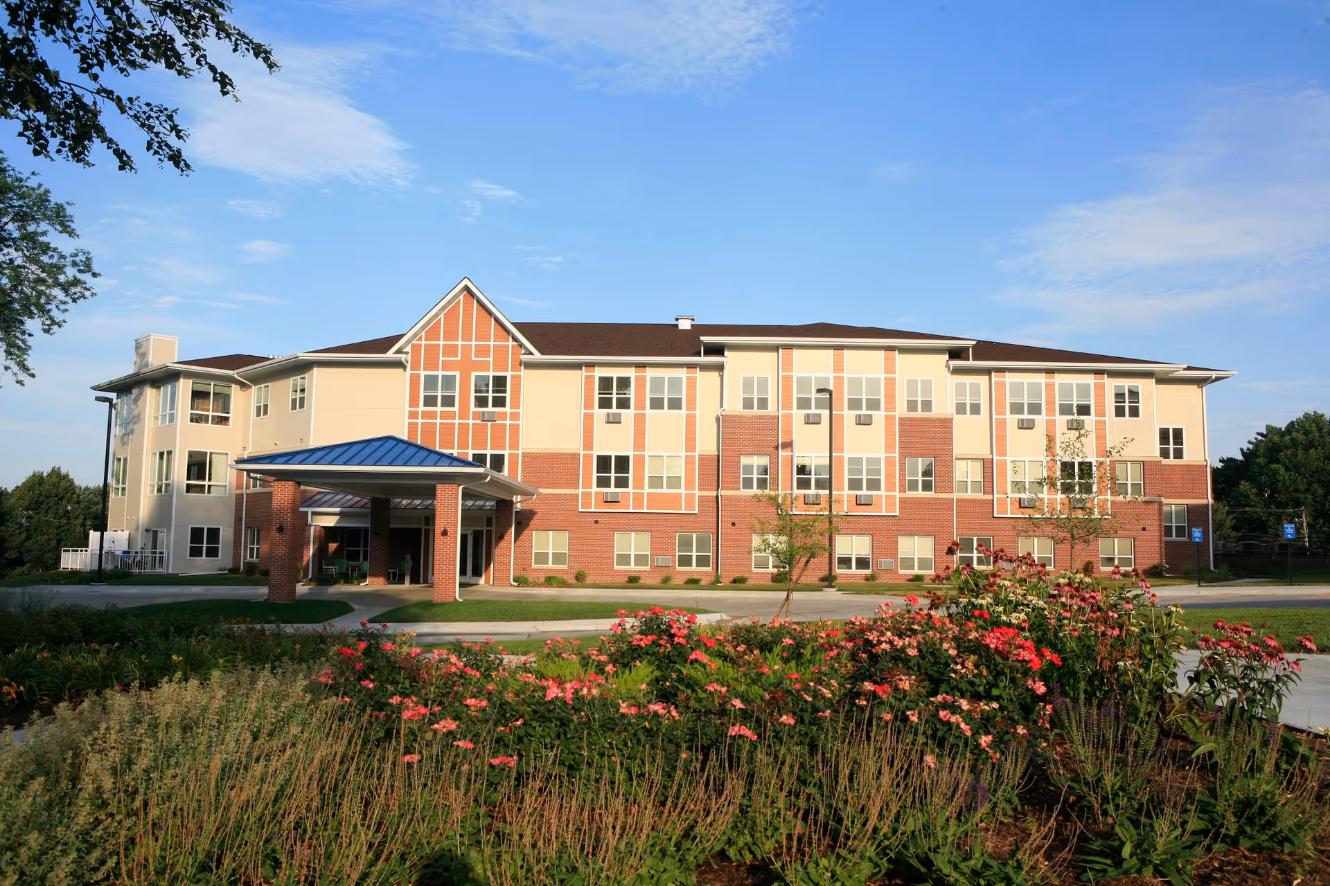 Exterior view of a three-story assisted living facility building with a covered entrance and a landscaped garden with flowers in the foreground under a clear blue sky.