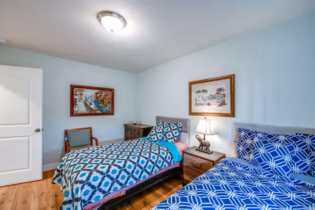A bedroom with two single beds, each with patterned blue bedding. Between the beds is a wooden nightstand with a lamp and a small elephant figurine. The room has light blue walls, wooden flooring, a wooden chair, and two framed pictures hanging on the walls.