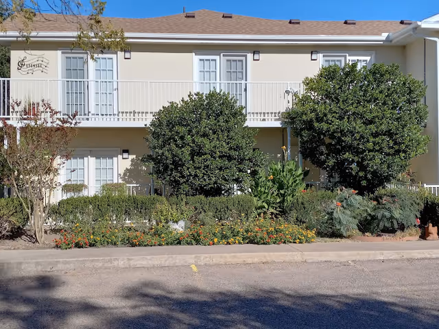 Exterior view of a two-story beige building with white railings and multiple windows. The building is surrounded by green bushes and colorful flowers in front, with a clear blue sky above.