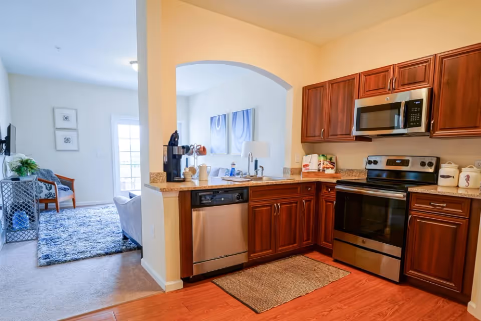 A modern kitchen with wooden cabinets, stainless steel appliances including a microwave, stove, and dishwasher. The kitchen has a granite countertop with a coffee maker and some decorative items. Adjacent to the kitchen is a living area with a blue rug, a chair, and a TV mounted on the wall. The space is well-lit with natural light coming through a glass door.