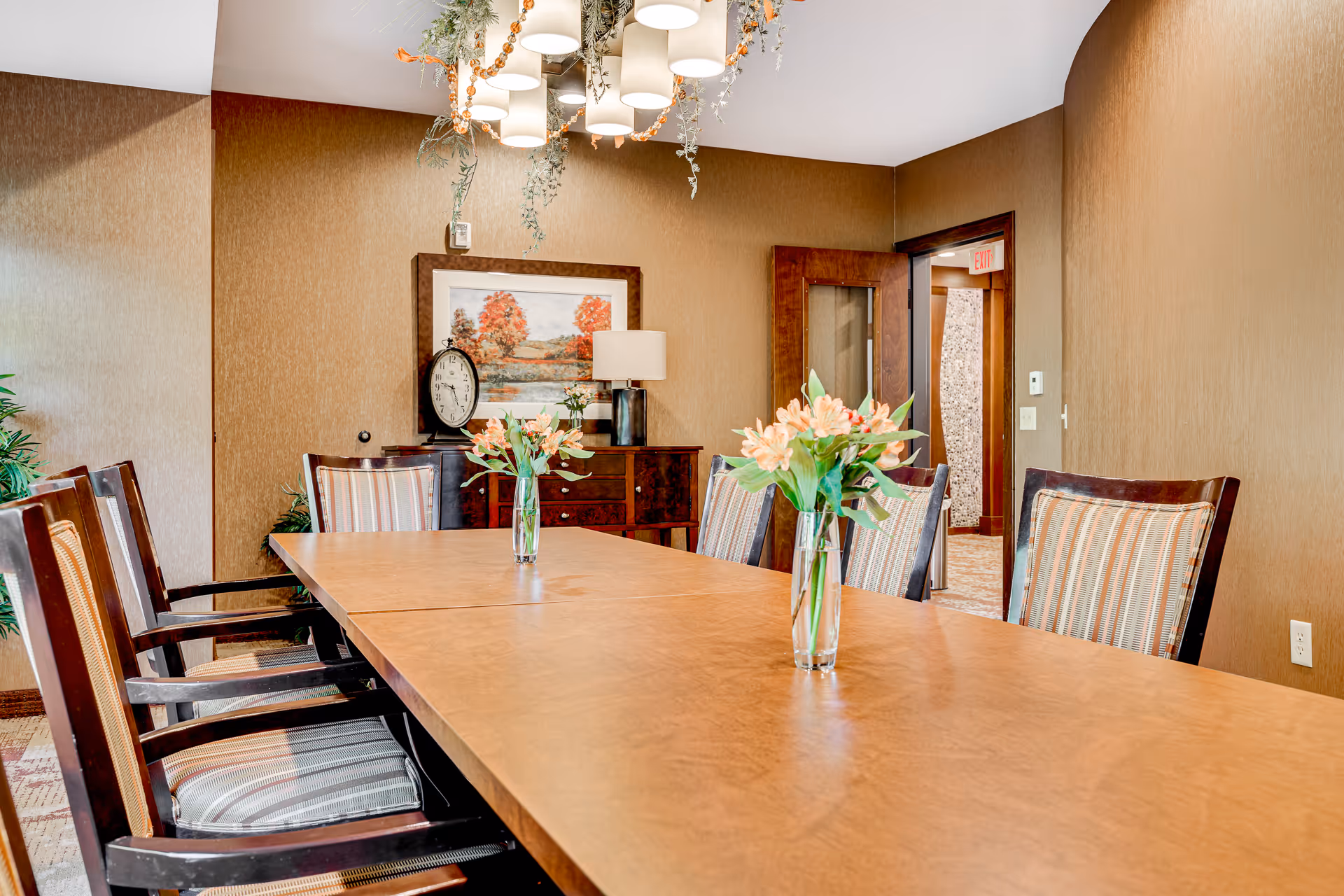 A dining room with a long wooden table surrounded by striped cushioned chairs. Two glass vases with flowers are placed on the table. The walls are brown, and there is a sideboard with a lamp, a clock, and a framed picture of autumn trees. A modern chandelier with hanging greenery is above the table.