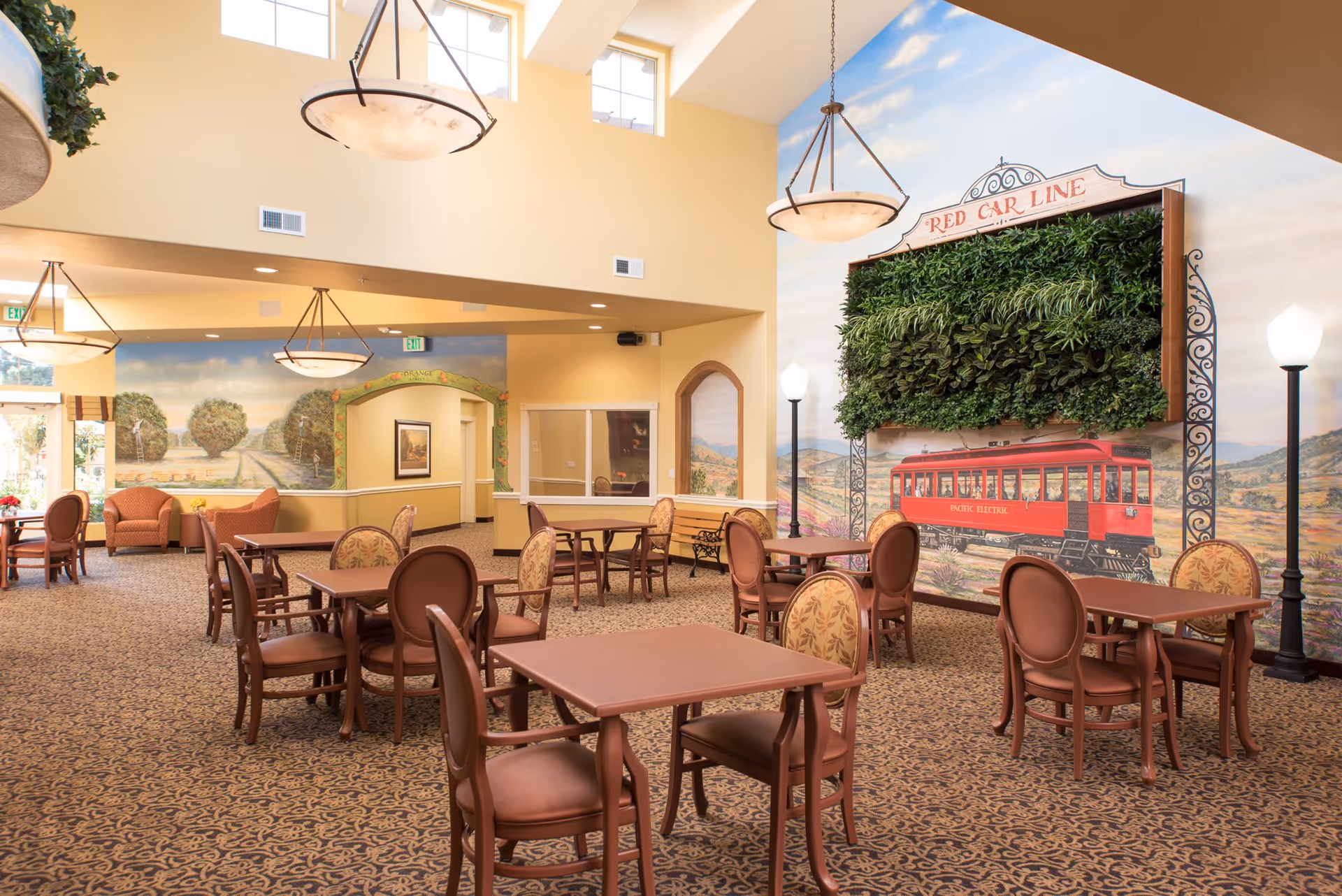 A spacious dining area with multiple wooden tables and chairs arranged on a patterned carpet. The walls are painted with murals depicting outdoor scenes, including a red trolley labeled 'Red Car Line' with greenery above it. The room has high ceilings with large hanging light fixtures and windows near the ceiling allowing natural light to enter.
