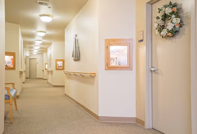 A well-lit hallway in a senior living facility with beige walls and carpeted floor. The hallway features handrails on both sides, framed display cases mounted on the walls, a wooden chair with a blue cushion, and a door decorated with a floral wreath.