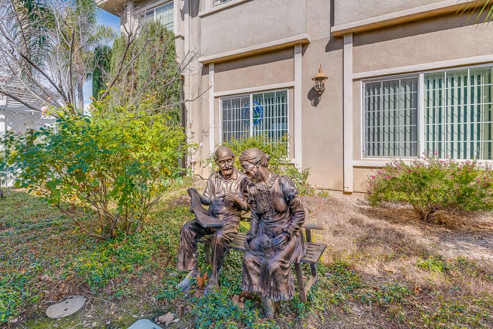 Bronze statues of an elderly man and woman sitting on a bench in a landscaped courtyard outside a senior living building.
