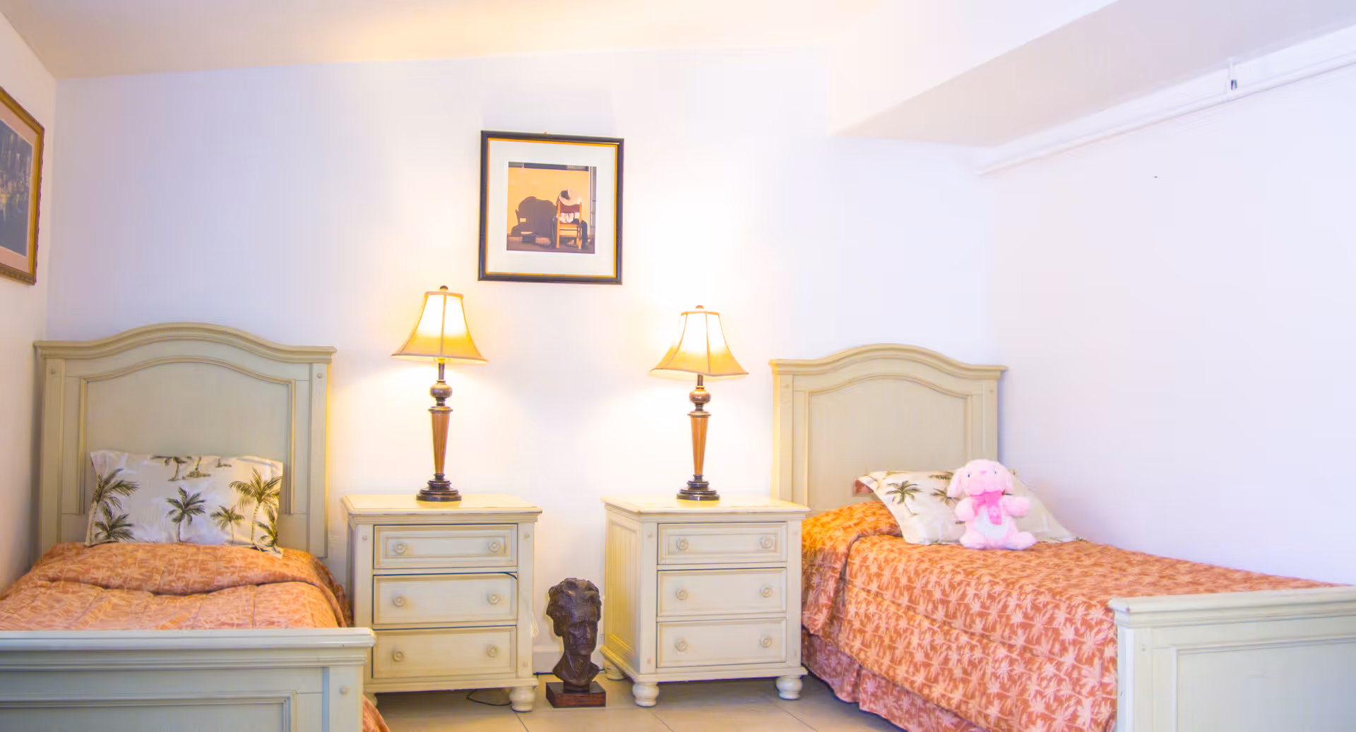 A bedroom with two single beds separated by two nightstands, each with a lamp. The beds have matching orange bedspreads and tropical-themed pillows. A pink stuffed animal is placed on the right bed. There are framed pictures on the walls above the beds and nightstands.