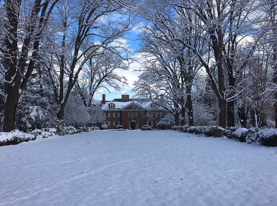 A large brick building in the distance surrounded by tall trees covered in snow, with a wide snow-covered lawn in the foreground under a partly cloudy blue sky.