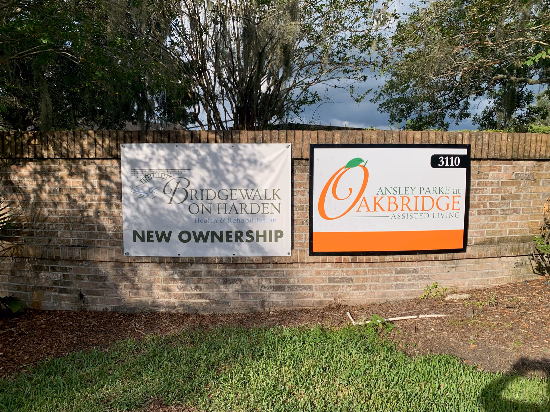 Two signs mounted on a brick wall outdoors. The left sign reads 'Bridgewalk on Harden Health & Rehabilitation NEW OWNERSHIP' with a small bridge graphic. The right sign reads '3110 Ansley Parke at Oakbridge Assisted Living' with an orange and green leaf graphic. Trees and grass are visible around the wall.