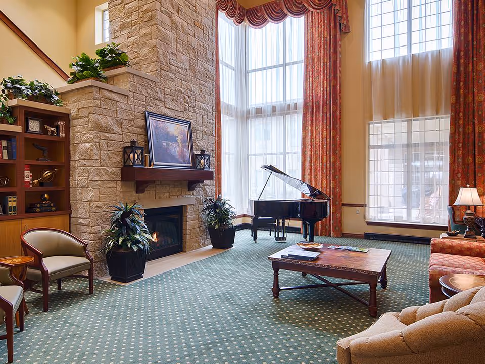 A spacious living room with a tall stone fireplace, a grand piano near large windows with red patterned curtains, a wooden coffee table, several upholstered chairs and sofas, and decorative plants and shelves.