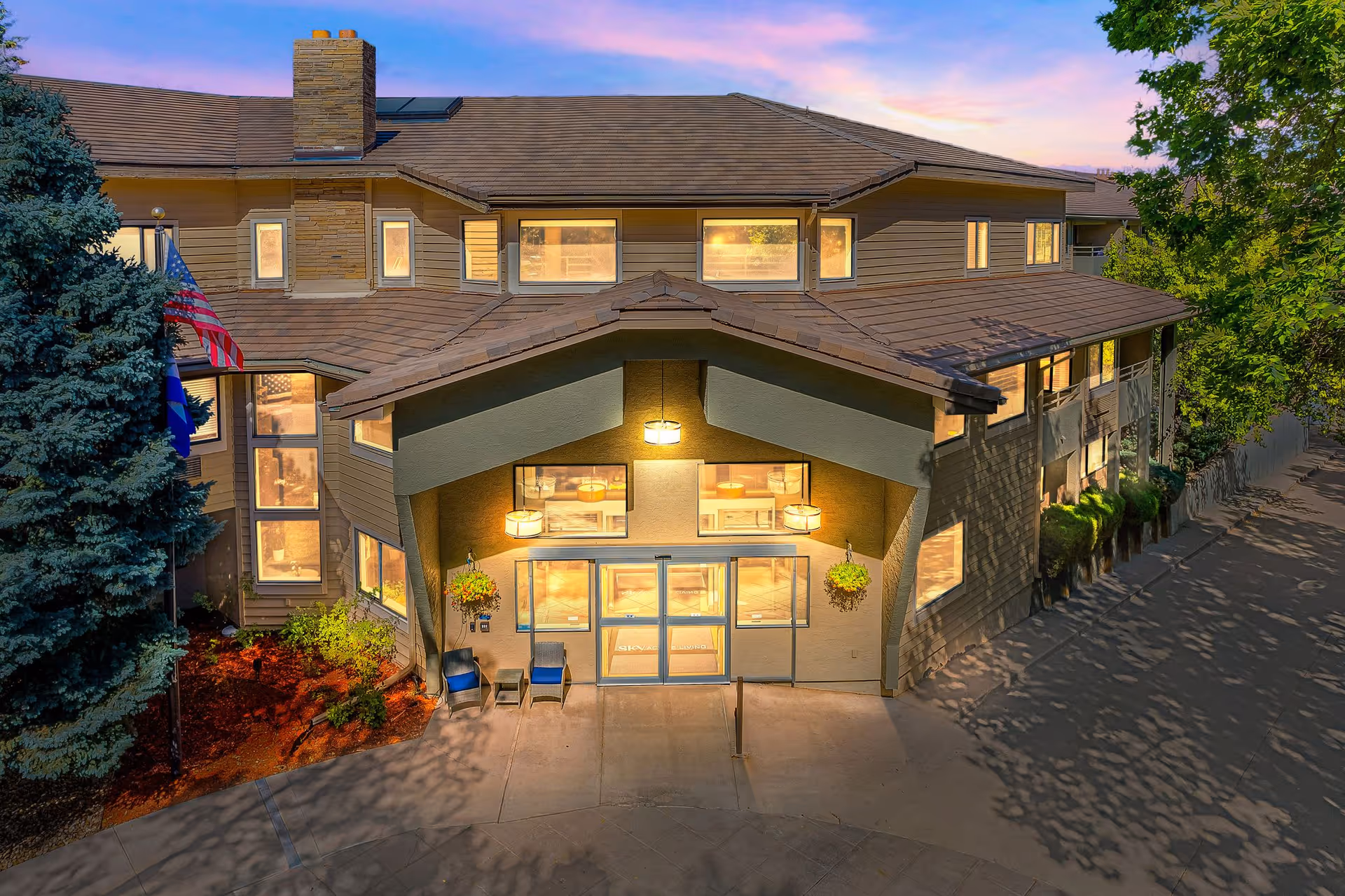 Exterior view of a two-story senior living facility building at dusk with warm lights glowing from the windows. The entrance features a covered porch with hanging plants and two chairs. There are trees and landscaping around the building.
