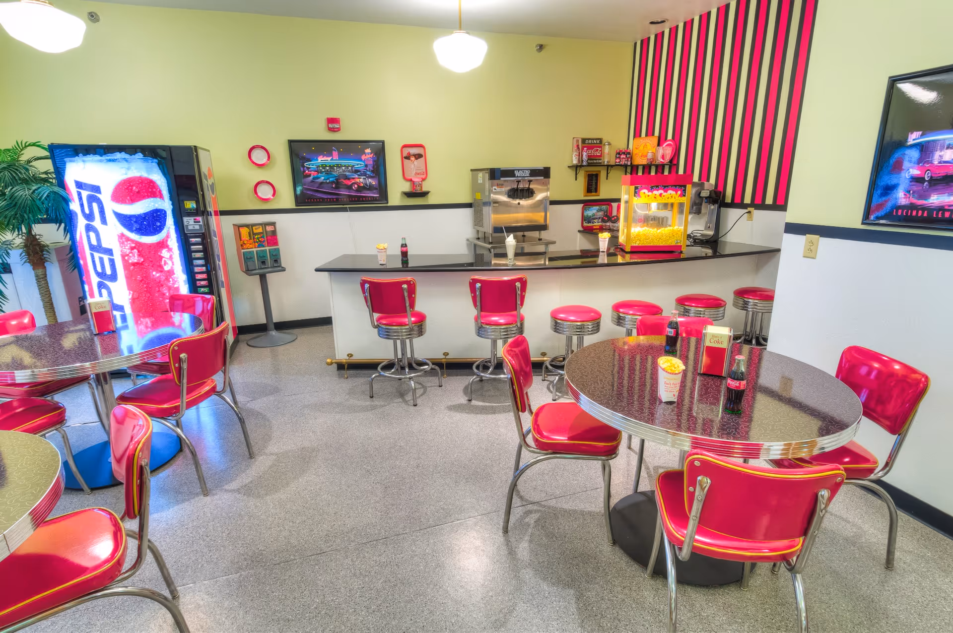 A retro-style dining area with bright pink chairs and stools around round tables and a counter. There is a Pepsi vending machine, a popcorn machine, and a milkshake machine on the counter. The walls are decorated with colorful artwork and striped wallpaper.